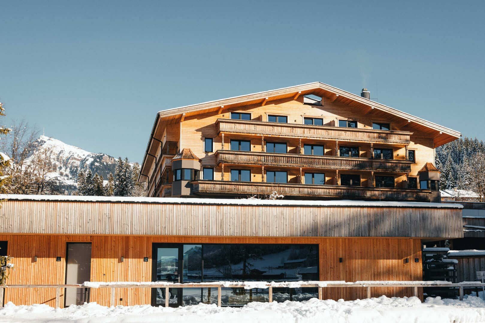 Alpenhotel Kitzbühel im Winter: vierstöckiges Holzhaus mit umlaufenden Balkonen, verschneiter Vorplatz, blauer Himmel