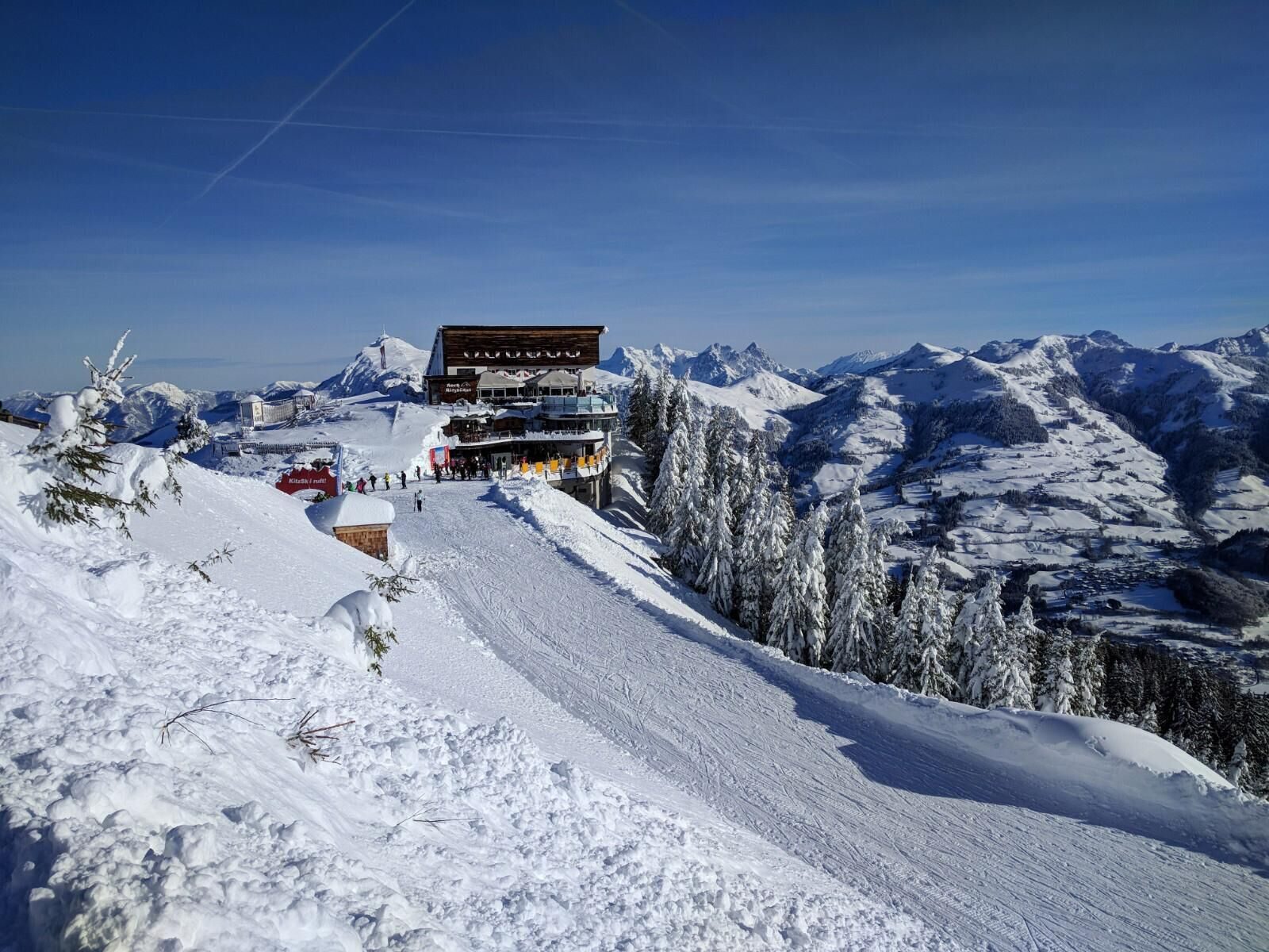 Holzbau des Hochkitzbühel an der Hahnenkamm-Bergstation, davor eine verschneite Piste mit Skifahrern, dahinter der Wilde Kaiser